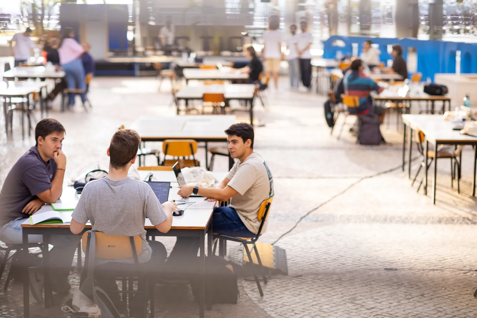 Three students working at laptops at a long table in a bright atrium